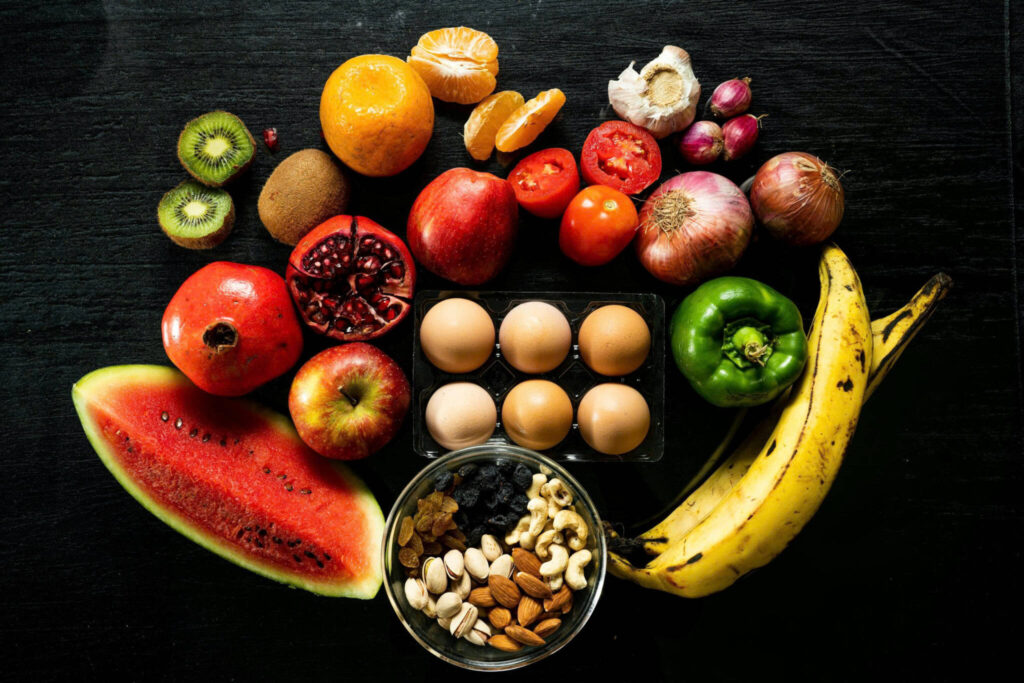 Overhead arrangement of fresh fruit, vegetables, eggs and nuts on a dark surface